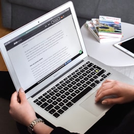 A person is using a MacBook Air, typing with both hands on the keyboard. The screen displays a document with text. In the background, a round white table holds a tablet and various books with colorful covers. The setting appears to be a comfortable home or office environment with a couch visible in the background.