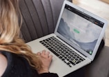 A person uses a MacBook Air laptop while sitting on a gray couch. The laptop screen displays a webpage with a serene mountain landscape and text related to remote jobs.