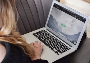 A person uses a MacBook Air laptop while sitting on a gray couch. The laptop screen displays a webpage with a serene mountain landscape and text related to remote jobs.