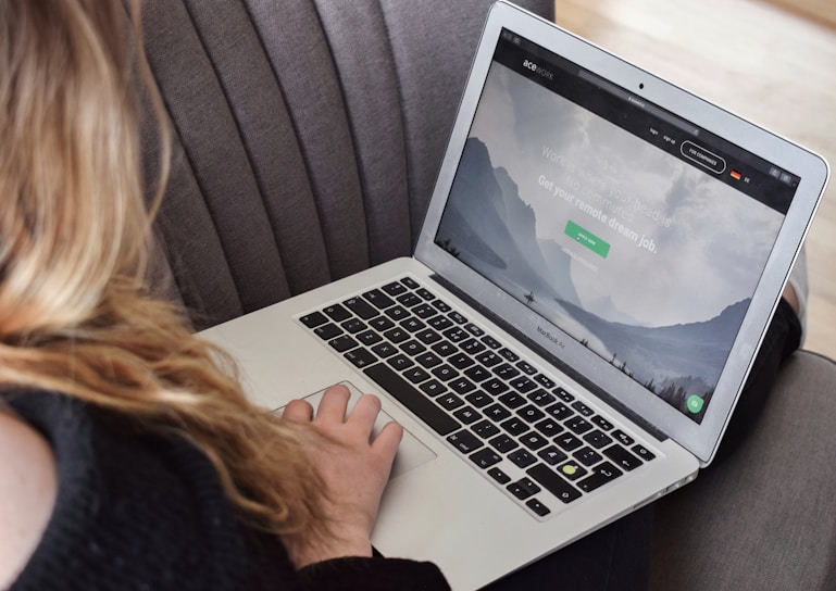A person uses a MacBook Air laptop while sitting on a gray couch. The laptop screen displays a webpage with a serene mountain landscape and text related to remote jobs.