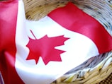 A Canadian flag with a red maple leaf in the center is draped inside a woven basket made from recycled newspaper. The flag's bright red and white colors contrast with the earthy tones of the basket.