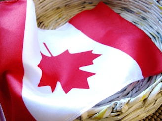 A Canadian flag with a red maple leaf in the center is draped inside a woven basket made from recycled newspaper. The flag's bright red and white colors contrast with the earthy tones of the basket.