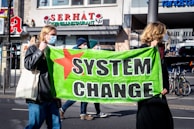 Two individuals wearing face masks are holding a bright green banner with the words 'SYSTEM CHANGE' and a red star on it. They appear to be participating in a street protest or demonstration. Other people and buildings are visible in the background, including a restaurant and what seems to be a pharmacy.