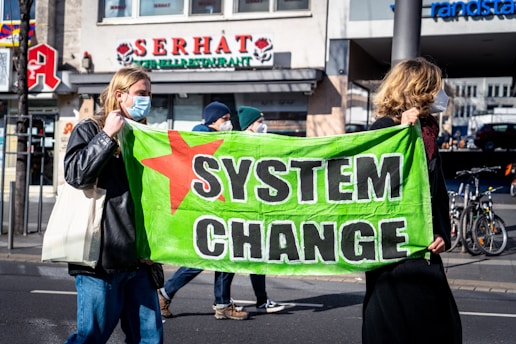 Two individuals wearing face masks are holding a bright green banner with the words 'SYSTEM CHANGE' and a red star on it. They appear to be participating in a street protest or demonstration. Other people and buildings are visible in the background, including a restaurant and what seems to be a pharmacy.
