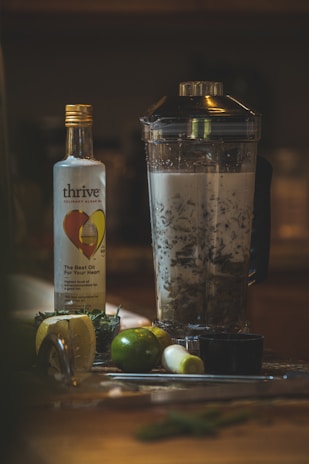A kitchen scene featuring a bottle of culinary algae oil with a heart on the label and a blender filled with murky liquid and leafy contents. A halved lemon, a whole lime, green onions, a small cup, and various herbs are scattered on the countertop.