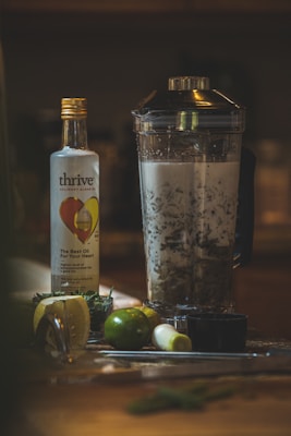 A kitchen scene featuring a bottle of culinary algae oil with a heart on the label and a blender filled with murky liquid and leafy contents. A halved lemon, a whole lime, green onions, a small cup, and various herbs are scattered on the countertop.