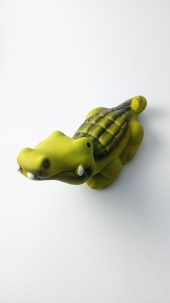 A small, cartoonish green crocodile toy with a rounded body and exaggerated features, resting on a light surface. The crocodile has a textured back, black eyes, and a smooth finish.