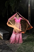 A person dressed in an elegant and colorful traditional outfit is standing outdoors, with large palm trees in the background. The attire consists of a vibrant pink top with intricate floral embroidery and a matching long skirt with a detailed pattern. A translucent scarf, bordered with an orange hue, is being gracefully held overhead. The scene is set against a dark backdrop, giving focus to the bright and intricately designed clothing.