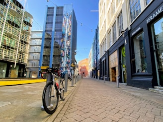 A bicycle is parked on an empty urban street surrounded by tall, modern buildings. A vibrant mural decorates one building with colorful patterns and figures. The street is paved with brick tiles and lit by string lights overhead.