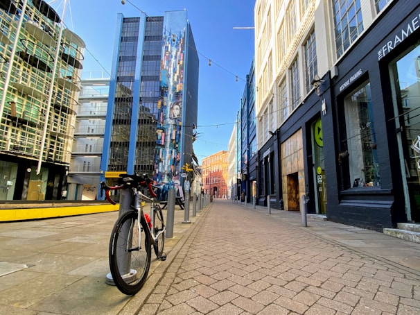 A bicycle is parked on an empty urban street surrounded by tall, modern buildings. A vibrant mural decorates one building with colorful patterns and figures. The street is paved with brick tiles and lit by string lights overhead.