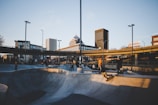 A bustling urban skatepark during the day, with several skateboarders actively engaging in skateboarding activities. The skatepark is surrounded by modern city architecture, including tall buildings and overpasses. The sunlight casts long shadows and warm hues across the scene, creating a dynamic atmosphere.