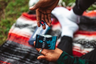 A person is holding a tea bag and its packaging over a vibrant, multicolored blanket. The tea bag is branded with a logo and the words 'Zest Tea'. The background shows grass, suggesting an outdoor setting.