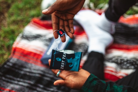 A person is holding a tea bag and its packaging over a vibrant, multicolored blanket. The tea bag is branded with a logo and the words 'Zest Tea'. The background shows grass, suggesting an outdoor setting.