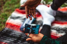 A person is holding a tea bag and its packaging over a vibrant, multicolored blanket. The tea bag is branded with a logo and the words 'Zest Tea'. The background shows grass, suggesting an outdoor setting.