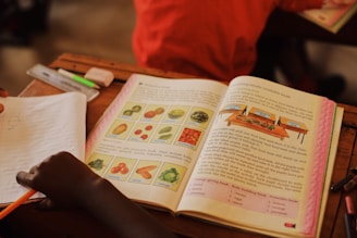 An open textbook on a desk features illustrations of various fruits and vegetables with labels, accompanied by detailed text. A child's hand is visible, holding a pencil over a notebook. Other items, such as a highlighter and eraser, are present on the desk.