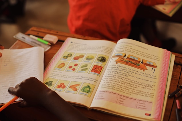 An open textbook on a desk features illustrations of various fruits and vegetables with labels, accompanied by detailed text. A child's hand is visible, holding a pencil over a notebook. Other items, such as a highlighter and eraser, are present on the desk.