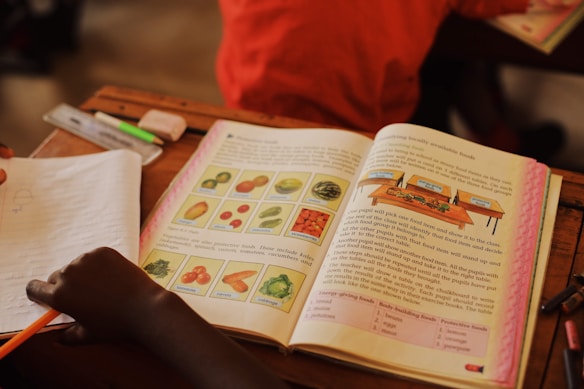 An open textbook on a desk features illustrations of various fruits and vegetables with labels, accompanied by detailed text. A child's hand is visible, holding a pencil over a notebook. Other items, such as a highlighter and eraser, are present on the desk.