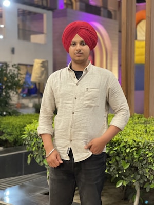 A person wearing a red turban and light-colored shirt stands in a landscaped area featuring green plants and modern architectural elements. The background includes soft lighting and colorful structures.