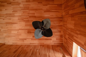 A ceiling fan with four wide, dark blades is mounted on a wooden ceiling with a rich texture. The room's walls and floor are composed of the same wood material, creating a warm and cohesive atmosphere. A window on the bottom right allows light to enter, contributing to the room's natural glow.