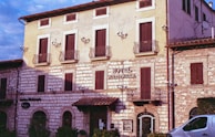 A historic building with stone walls and wooden shutters, identified as a hotel. The facade features balconies with iron railings and decorative wall lamps. A small car is partially visible in the foreground, parked near the building.