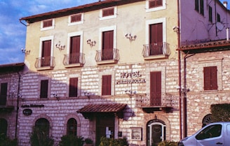 A historic building with stone walls and wooden shutters, identified as a hotel. The facade features balconies with iron railings and decorative wall lamps. A small car is partially visible in the foreground, parked near the building.