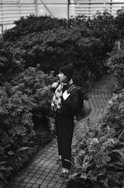 A person is walking along a brick path surrounded by lush greenery. The individual is wearing a hat, scarf, and has a backpack, appearing deep in thought or observation.