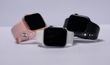 Three smartwatches are displayed on a light background. They feature different colored bands: pink, white, and black. The watches have large, rectangular touchscreens, and they are positioned to show off their sleek and modern design.