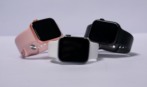 Three smartwatches are displayed on a light background. They feature different colored bands: pink, white, and black. The watches have large, rectangular touchscreens, and they are positioned to show off their sleek and modern design.