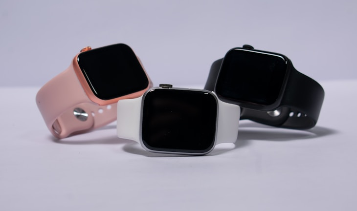 Three smartwatches are displayed on a light background. They feature different colored bands: pink, white, and black. The watches have large, rectangular touchscreens, and they are positioned to show off their sleek and modern design.