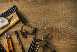 A wooden surface is scattered with various hand tools and hardware items. There is a hammer with a wooden handle, a black metal ruler, several screws, a cloth, pliers, drill bits, hex keys, and a utility knife with an orange and black casing.