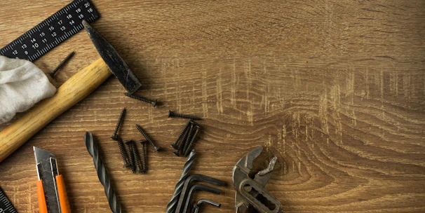 A wooden surface is scattered with various hand tools and hardware items. There is a hammer with a wooden handle, a black metal ruler, several screws, a cloth, pliers, drill bits, hex keys, and a utility knife with an orange and black casing.