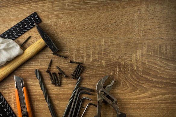 A wooden surface is scattered with various hand tools and hardware items. There is a hammer with a wooden handle, a black metal ruler, several screws, a cloth, pliers, drill bits, hex keys, and a utility knife with an orange and black casing.