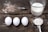 A rustic wooden surface holds three white eggs aligned horizontally next to a whisk with some scattered flour. Nearby, a measuring cup is filled with milk, and a metal measuring spoon holds flour, suggesting preparation for baking.