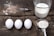 A rustic wooden surface holds three white eggs aligned horizontally next to a whisk with some scattered flour. Nearby, a measuring cup is filled with milk, and a metal measuring spoon holds flour, suggesting preparation for baking.