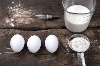 A rustic wooden surface holds three white eggs aligned horizontally next to a whisk with some scattered flour. Nearby, a measuring cup is filled with milk, and a metal measuring spoon holds flour, suggesting preparation for baking.