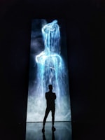 A silhouette of a person stands in front of a tall, vertical digital display showcasing vibrant blue and white light resembling a waterfall or abstract liquid flow against a dark background.