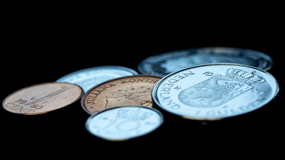 A collection of various coins is arranged at different angles on a dark surface. The coins vary in size and color, with some appearing silver and others showing a bronze hue. Intricate designs and inscriptions are visible, including recognizable figures and text.