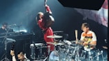 A live music performance featuring a musician energetically playing an upright bass, another drummer in a white and orange shirt playing drums, and a person playing the keyboard. The stage lighting creates a vibrant atmosphere, and there are various musical instruments visible, including a microphone and drum set.