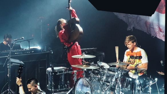 A live music performance featuring a musician energetically playing an upright bass, another drummer in a white and orange shirt playing drums, and a person playing the keyboard. The stage lighting creates a vibrant atmosphere, and there are various musical instruments visible, including a microphone and drum set.