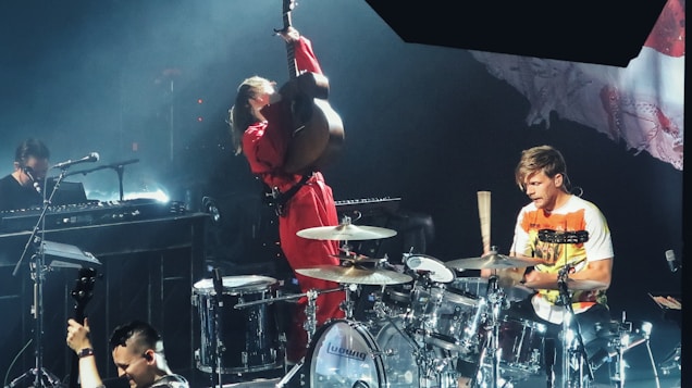 A live music performance featuring a musician energetically playing an upright bass, another drummer in a white and orange shirt playing drums, and a person playing the keyboard. The stage lighting creates a vibrant atmosphere, and there are various musical instruments visible, including a microphone and drum set.