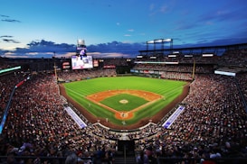 A baseball stadium filled with spectators. The field is brightly lit, with players positioned on the grass. The sky is a deep blue, dotted with clouds, hinting at an evening or late afternoon setting. Large screens and billboards display advertisements and game-related content.