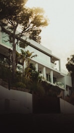 A modern white multi-level house surrounded by lush greenery with tall trees and various plants in the foreground. The design features large glass windows and clean architectural lines. The lighting appears to be natural with a soft, atmospheric glow.