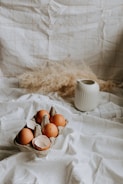 A small carton containing brown eggs is placed on a white cloth. One of the eggs is cracked, revealing its broken shell. Behind the carton is a white ceramic pitcher and a light, textured fabric backdrop with a grid pattern. The scene has a rustic and natural feel with neutral tones.