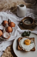 A breakfast setting featuring a piece of toast with a fried egg on top and garnished with fresh parsley. Next to it is a carton containing several brown eggs, some of which are cracked open. A cup of coffee sits on a saucer nearby, along with a white ceramic jug on a textured fabric background.