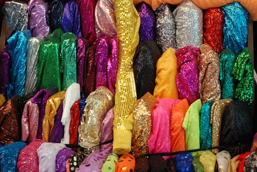 A colorful and vibrant display of folded fabric rolls, featuring numerous shades and textures, including shiny and sequined materials. The arrangement showcases a wide variety of colors such as pink, green, blue, gold, and more, creating a visually striking pattern.