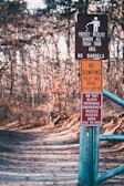 A forest path is lined with several warning signs attached to a metal post. The signs include messages about protecting wildlife, prohibiting dumping, and keeping the entrance clear for emergency vehicles. The path is wide, covered with leaves, and surrounded by bare trees, indicating a winter or early spring setting.
