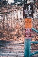 A forest path is lined with several warning signs attached to a metal post. The signs include messages about protecting wildlife, prohibiting dumping, and keeping the entrance clear for emergency vehicles. The path is wide, covered with leaves, and surrounded by bare trees, indicating a winter or early spring setting.