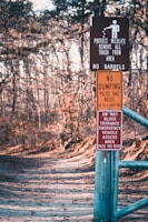 A forest path is lined with several warning signs attached to a metal post. The signs include messages about protecting wildlife, prohibiting dumping, and keeping the entrance clear for emergency vehicles. The path is wide, covered with leaves, and surrounded by bare trees, indicating a winter or early spring setting.