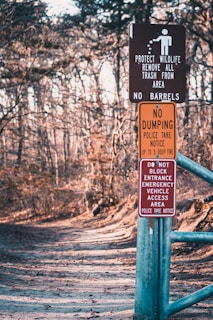 A forest path is lined with several warning signs attached to a metal post. The signs include messages about protecting wildlife, prohibiting dumping, and keeping the entrance clear for emergency vehicles. The path is wide, covered with leaves, and surrounded by bare trees, indicating a winter or early spring setting.