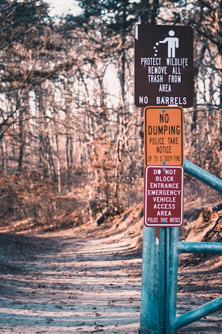 A forest path is lined with several warning signs attached to a metal post. The signs include messages about protecting wildlife, prohibiting dumping, and keeping the entrance clear for emergency vehicles. The path is wide, covered with leaves, and surrounded by bare trees, indicating a winter or early spring setting.